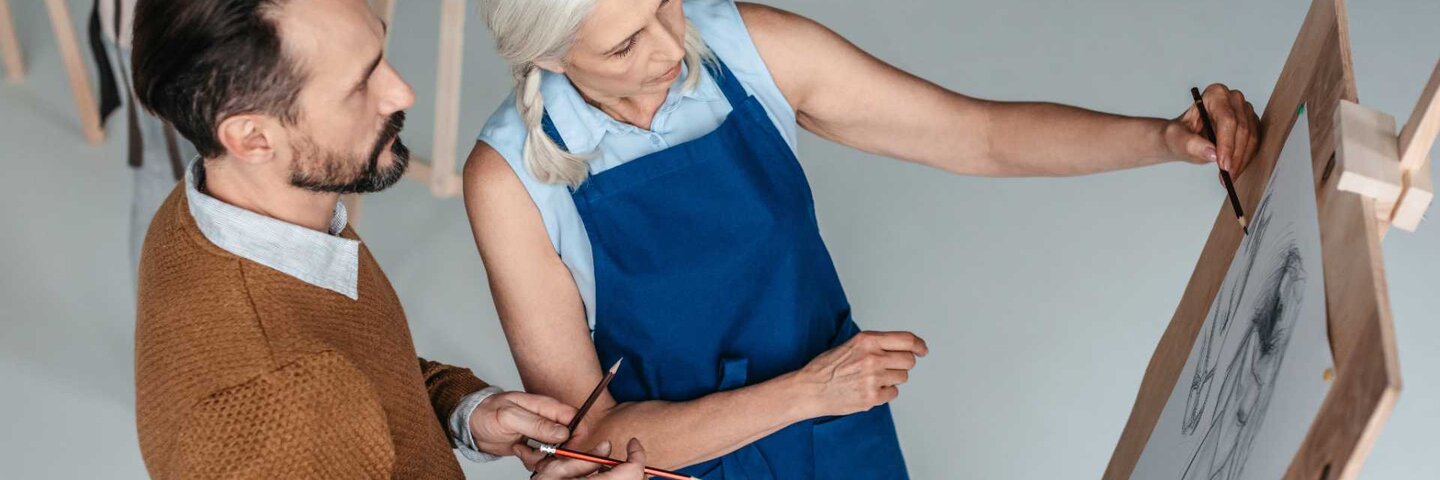 A mature man holding pencils and looking at senior woman drawing on easel