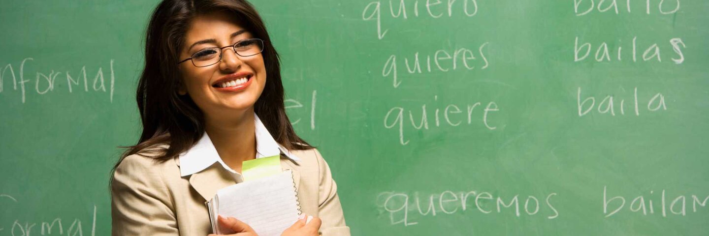 Teacher in classroom with spanish verbs written on board.